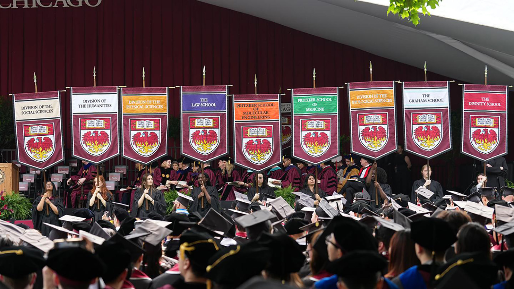 Students lined up before a stage at UChicago displaying colorful banners of the various schools and divisions