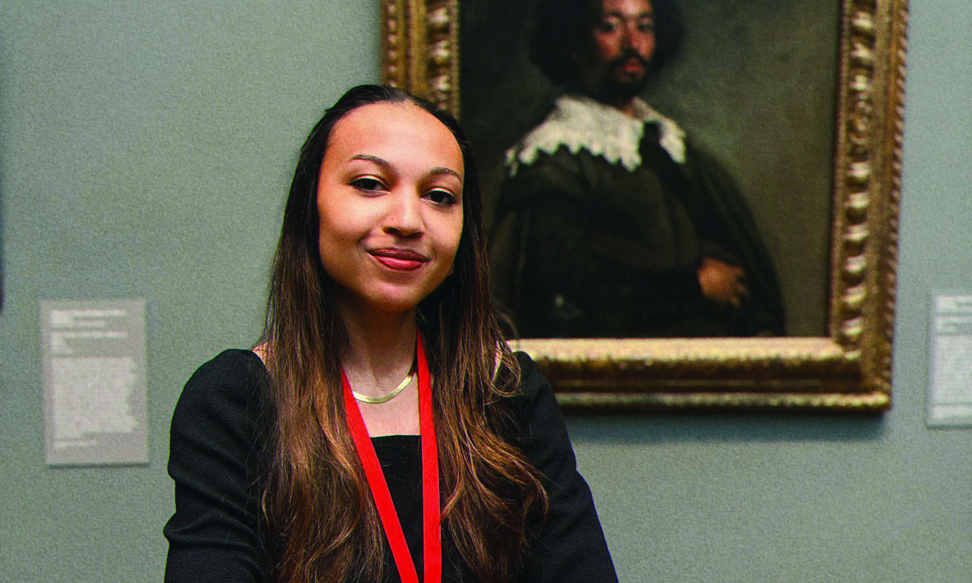 Portrait of Lela Jenkins looking directly at the camera while standing before a painting in an art museum wearing a black shirt and a red lanyard