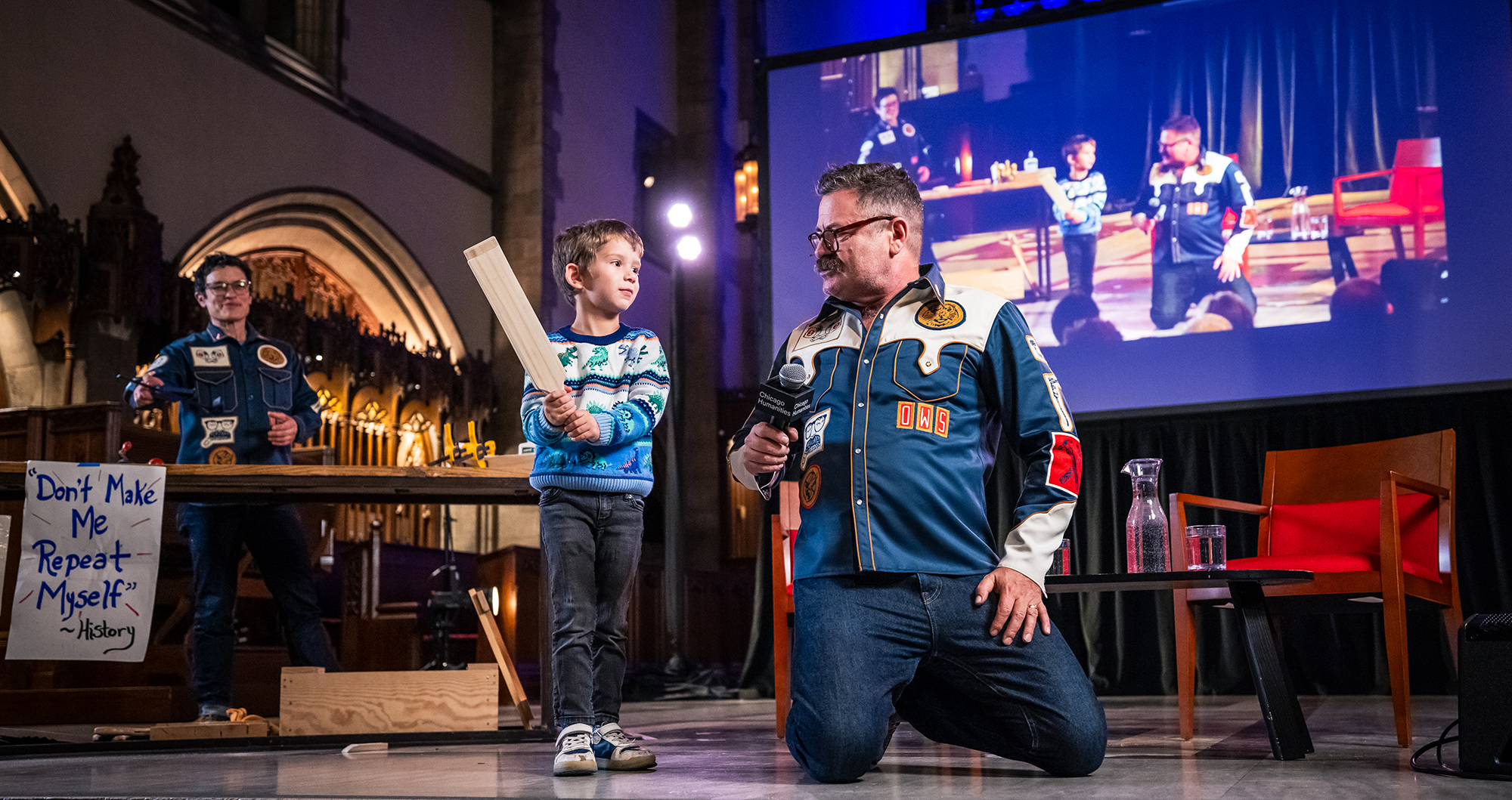 Nick Offerman wearing a dark blue shirt with patches kneels next to a child in a blue patterned sweater who is holding a large wooden slap stick.