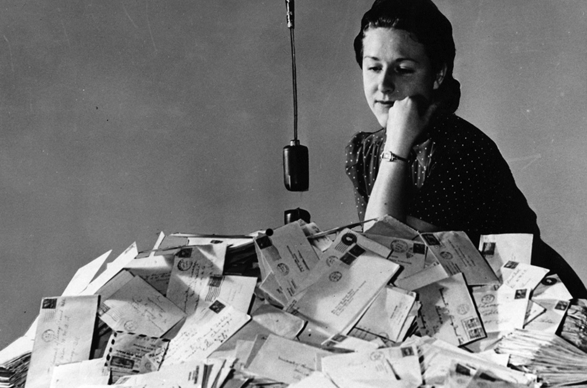 This is a black and white photograph showing a person seated behind a very large pile of mail, mostly envelopes. A hanging microphone is positioned in front of them. The overall style and clothing suggest a mid 20th century time period.