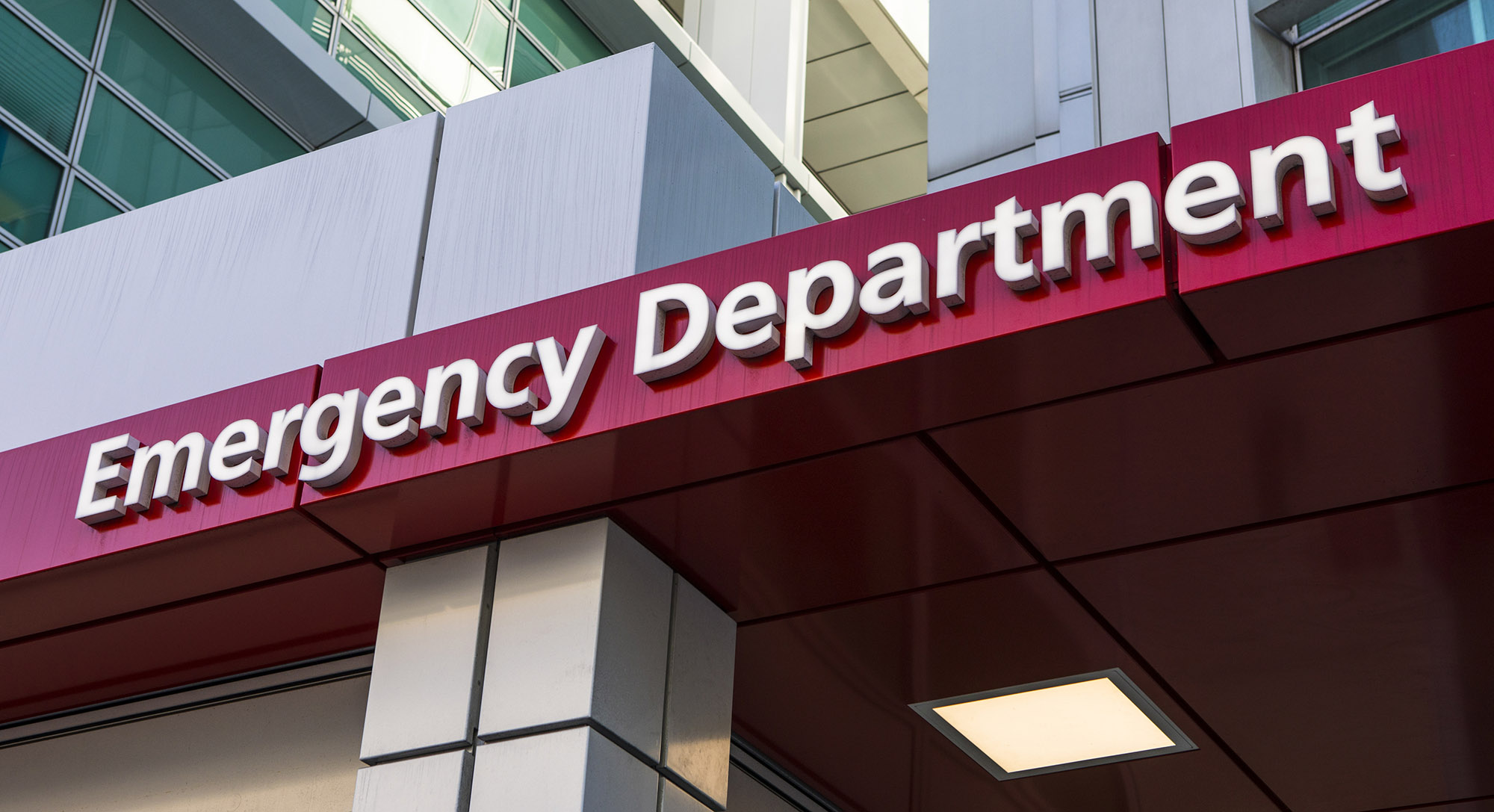 The image shows the entrance canopy of a hospital’s Emergency Department, with large white letters on a red background mounted on the building exterior.