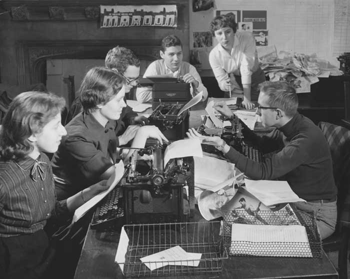 A group of college students in the 1950s sitting around a table with several manual typewriters and lots of paper 