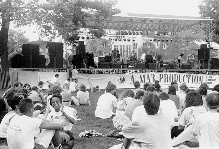 Students sit in the grass in front of an outdoor stage where musicians prepare to perform. A banner reads "A MAB Production Major Activities Board"
