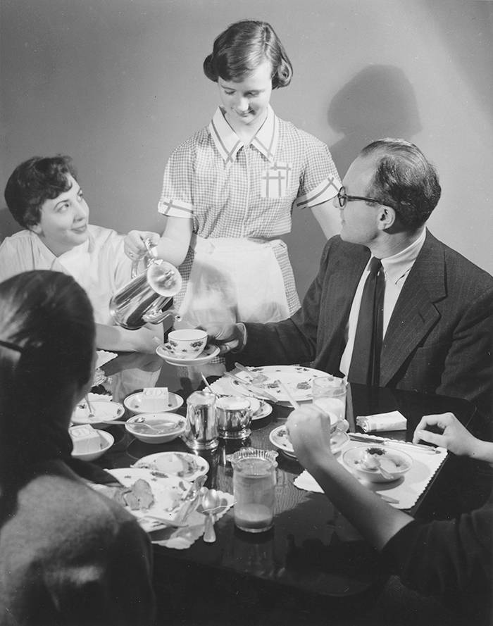 A young woman in an apron pours coffee for a man seated at a table with other people