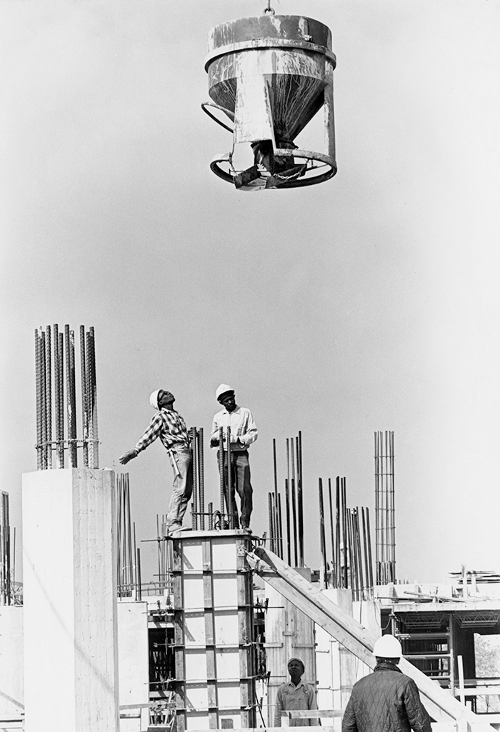 A cement mixer is lowered into a construction zone. Four construction workers wait to receive it