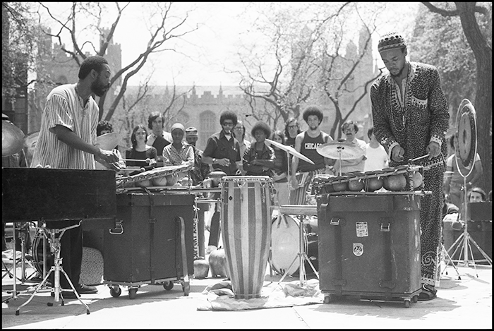Two percussionists play for a group of students. Gothic campus building and trees in the background