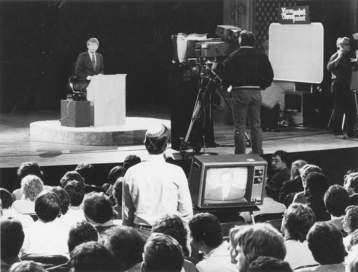 Man in suit sits on stage in front of a large video camera. An audience member stands to ask a question
