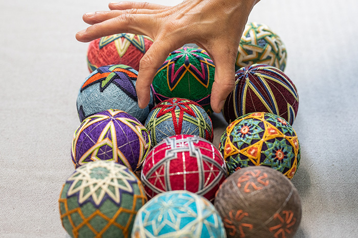 Hand reaches down toward colorfully embroidered temari balls, arranged in rows