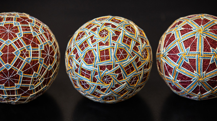 Three temari balls, embroidered with geometric patterns in maroon, blue, yellow, and white thread