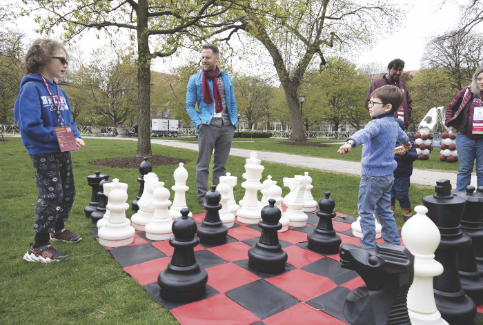 Children playing a giant chess game outside while adults look on.