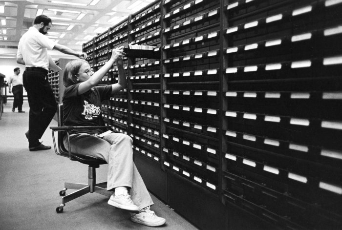 A B&W photo of a young woman sitting in a chair pulling out a drawer in a card catalog.