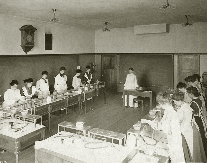 Black and white photo, about a dozen women stand around tables in a classroom, a woman stands in the center of the room in front of a chalkboard, next to a desk