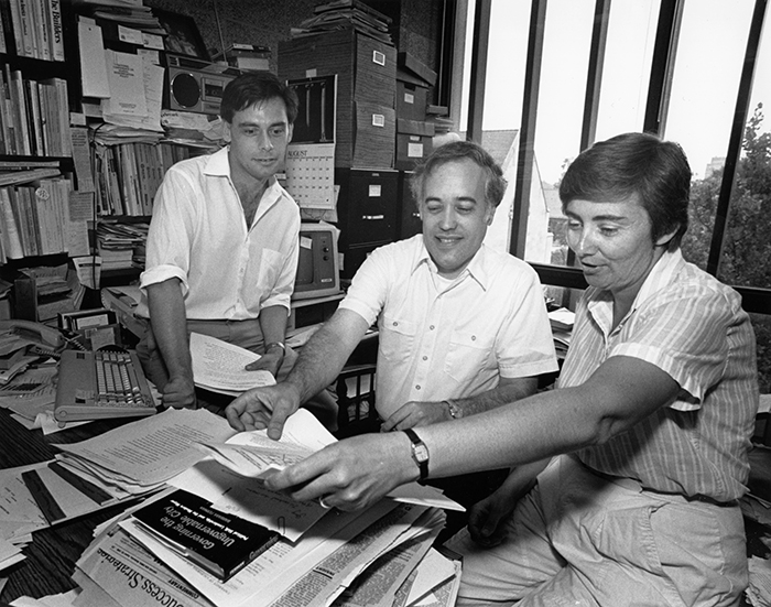 Three people sit at a cluttered desk and look at a piece of paper. In the background are bookshelves, file cabinets with boxes piled on top, and a wall of windows.