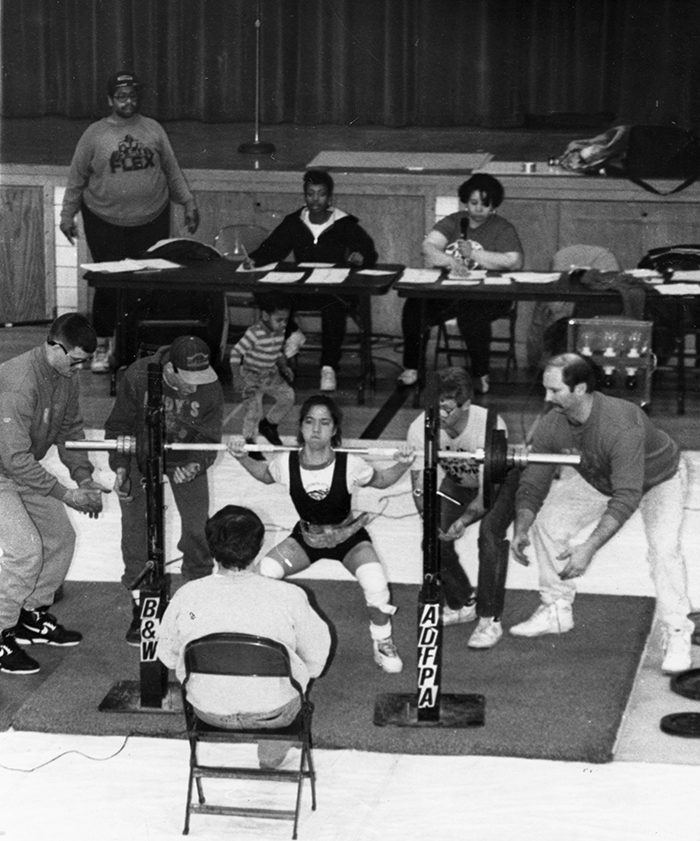 A young woman completes a squat, spotted by four men. Judges sit at a long table in the background with papers spread in front of them.