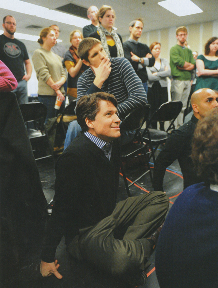 A group of about 15 people sit on the floor, folding chairs, or stand. They look toward the right.