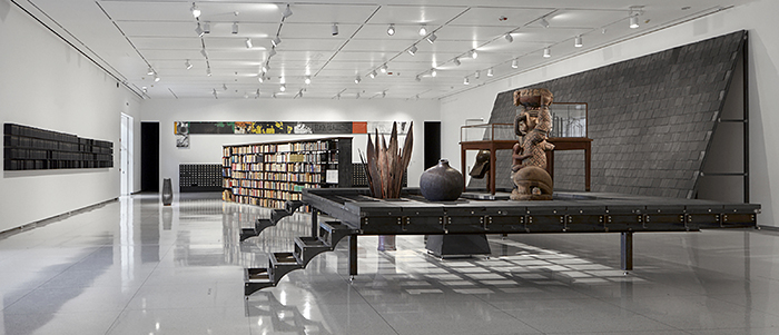 This image shows a contemporary art museum installation that combines sculpture, architectural elements, books, and archival lantern slides in original cabinets.