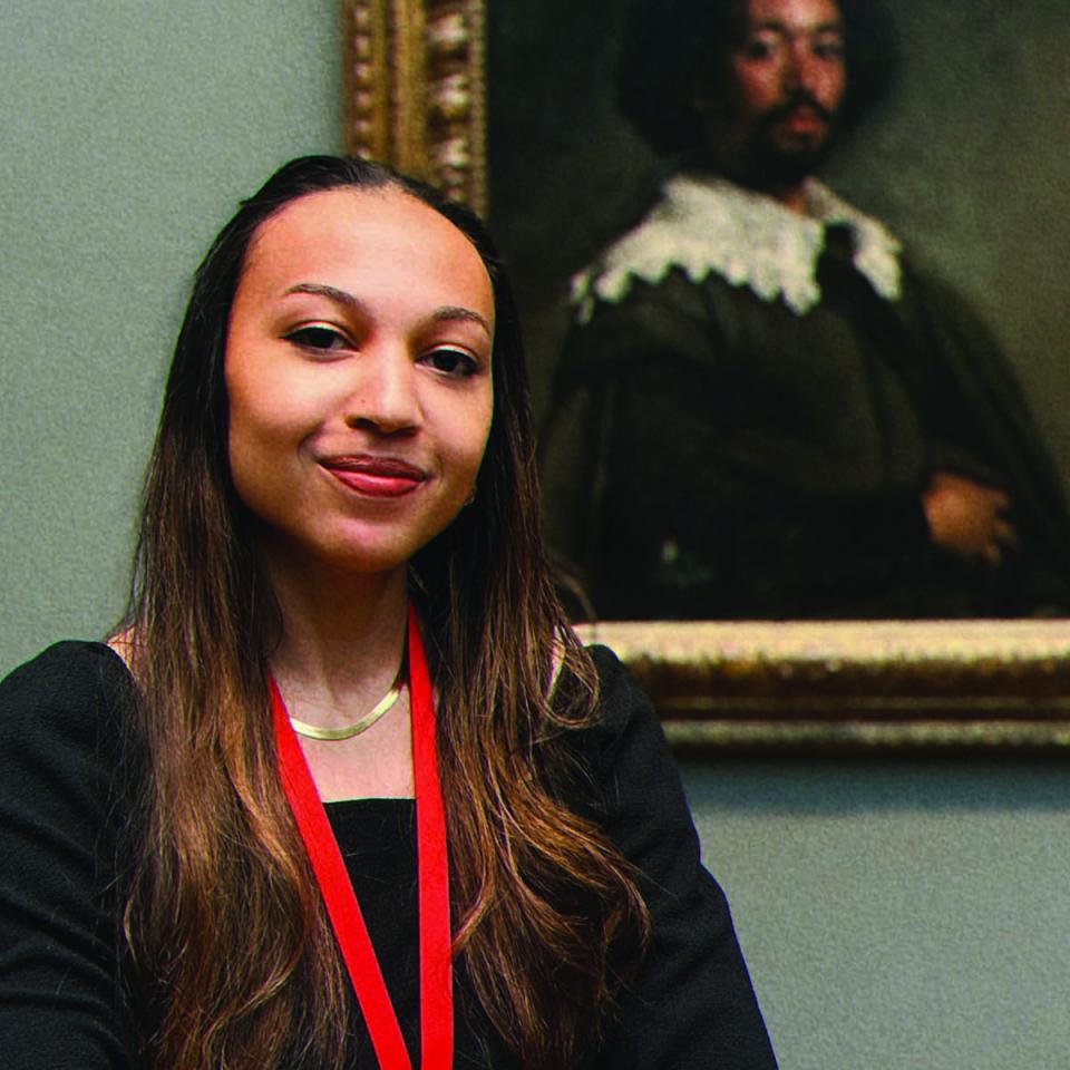 Portrait of Lela Jenkins looking directly at the camera while standing before a painting in an art museum wearing a black shirt and a red lanyard