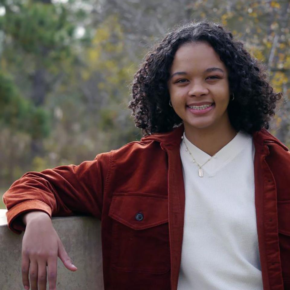 A person wearing a rust-colored jacket with large buttons and front pockets stands outdoors leaning casually against a light-colored stone or concrete structure with one arm resting on it.