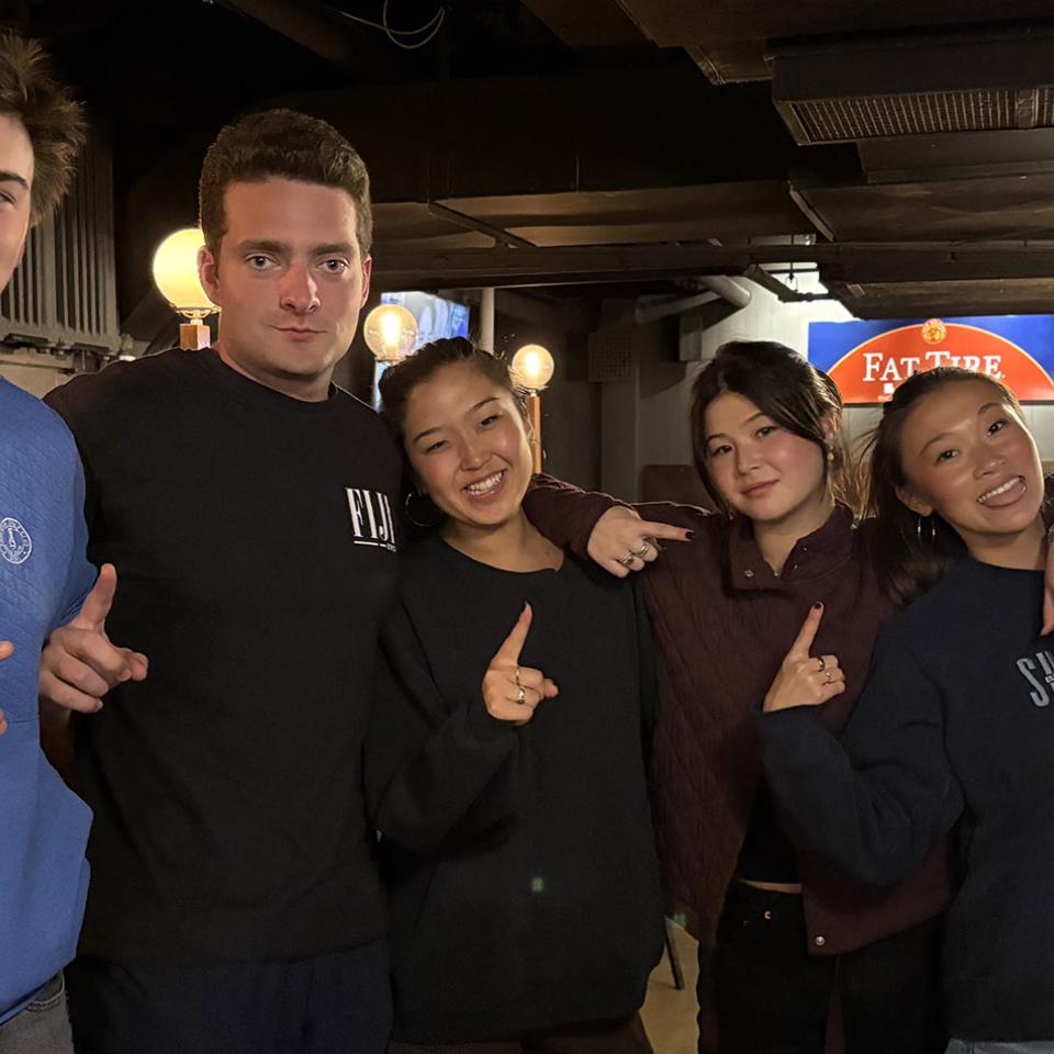 This photo shows a group of seven people standing closely together in a pub. They’re all facing the camera with their arms around each other’s shoulders, and each person is holding up one finger to celebrate their trivia win.